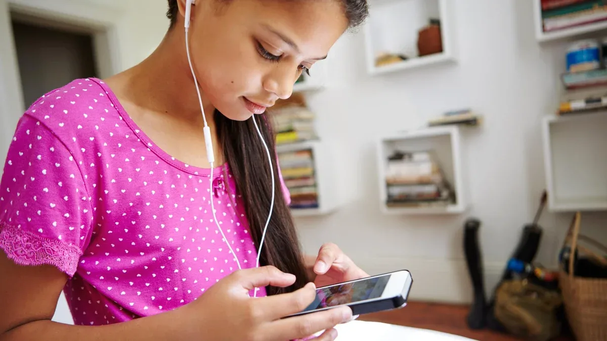 Adolescente tapant sur son téléphone dans sa chambre, écoutant de la musique, des podcasts et de l'audio en ligne avec des écouteurs. Sur son lit, elle gère une playlist via les réseaux sociaux pour son abonnement de streaming et écoute un album.